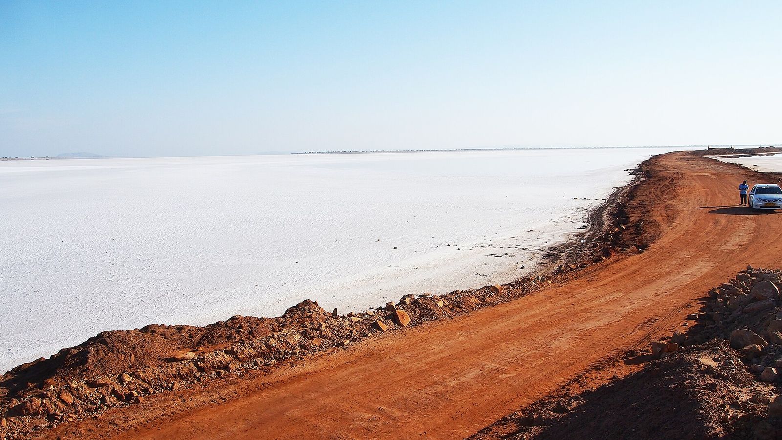 A red dirt road running beside the pale white salt flats of the Rann of Kutch, with a car parked at the edge
