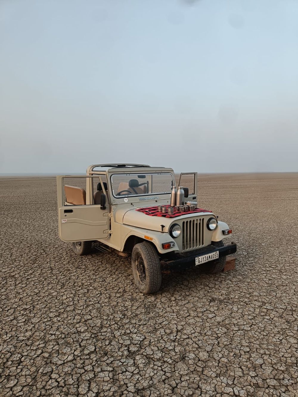 A cream Mahindra jeep with its doors open on a wide plain of cracked dry earth, tea flasks and cups set out on a red checked cloth on the bonnet