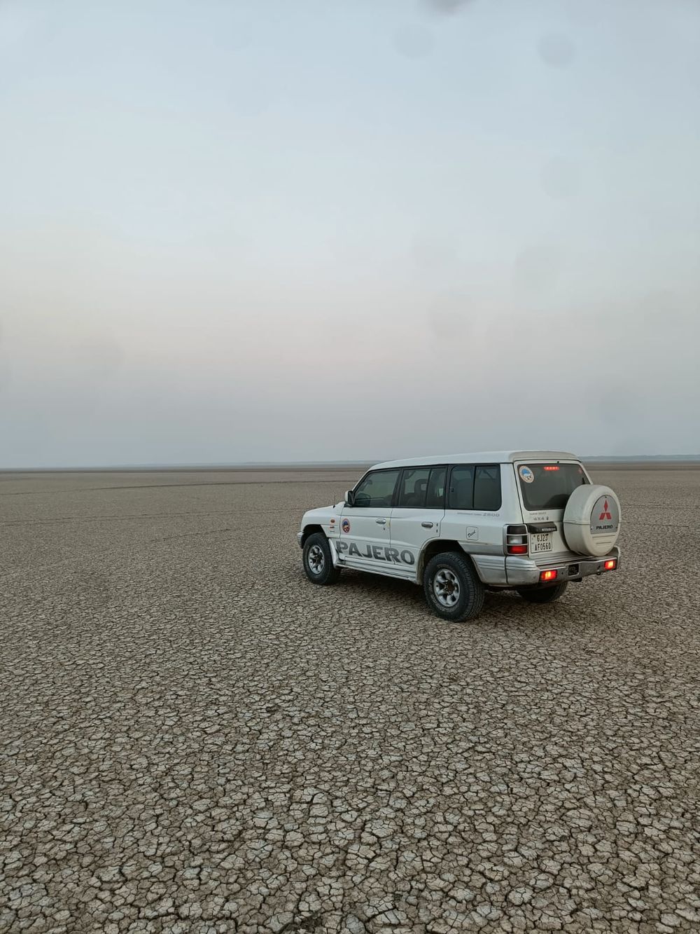 A white Mitsubishi Pajero seen from behind on a flat plain of cracked dry earth under a hazy sky
